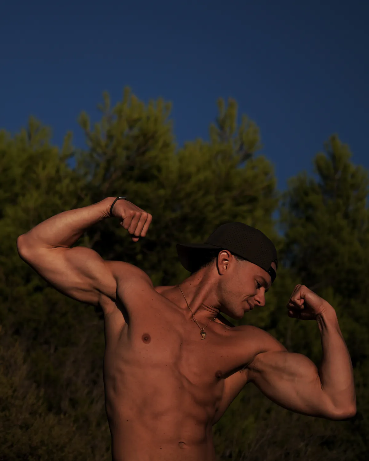 Paul torse nu et casquette, qui contracte les deux biceps en souriant, devant des pins et un ciel bleu profond.