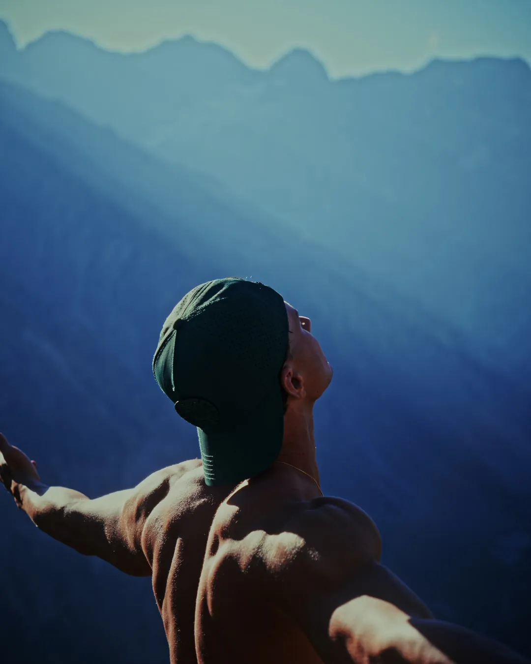 Paul de dos, torse nu et casquette, les bras ouverts et la tête levée vers le ciel, face à des montagnes bleutées.