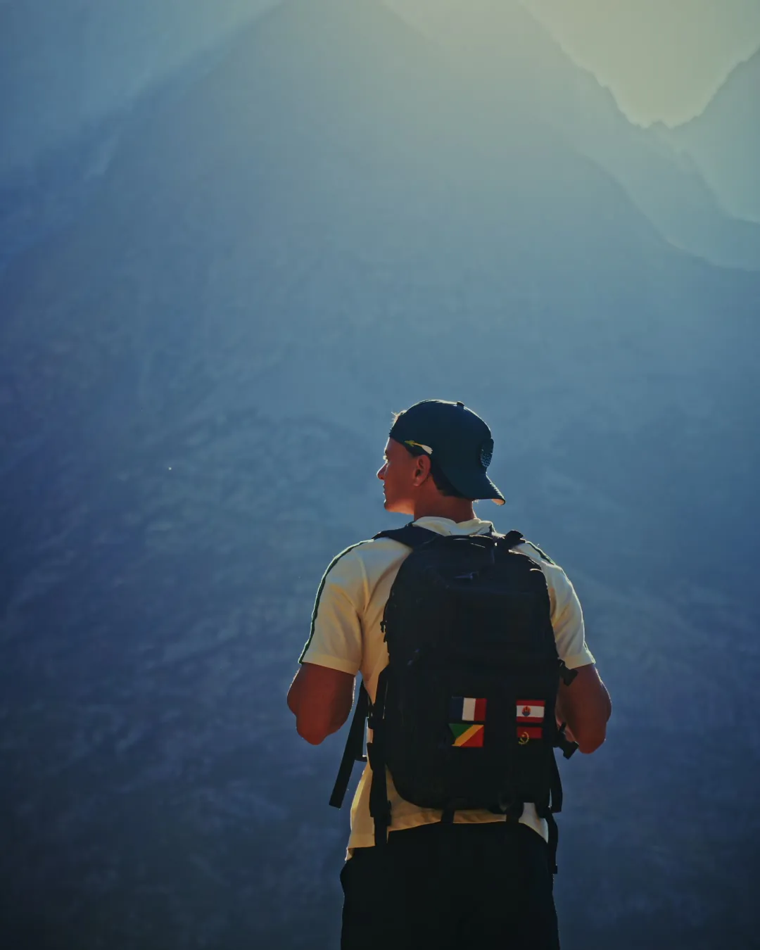 Paul de dos, casquette et sac à dos, face à une grande montagne dans une lumière bleutée.