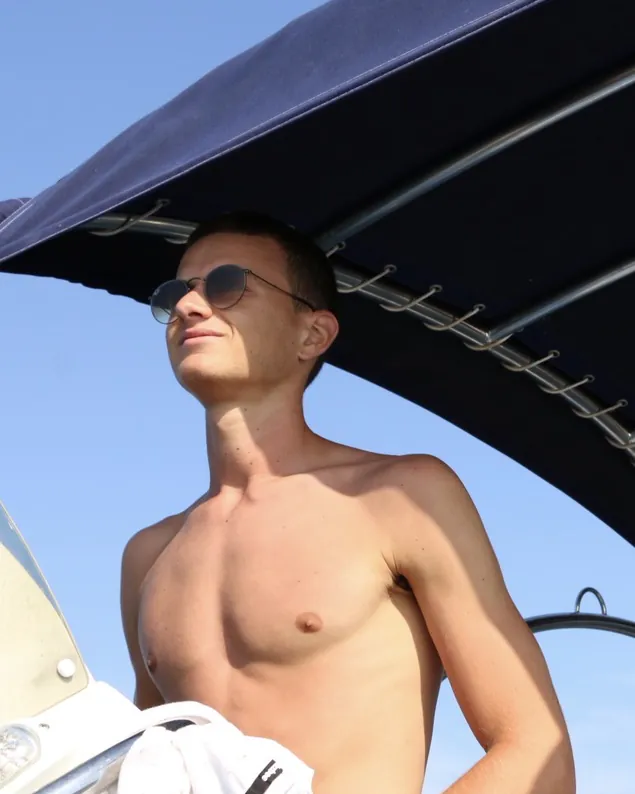 Paul, torse nu et lunettes de soleil, debout à l'avant d'un bateau, le regard au loin.