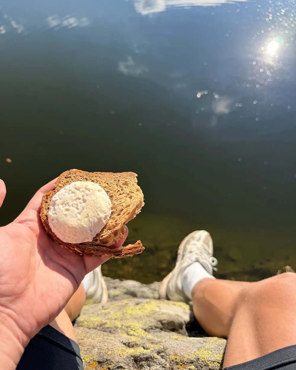 Une tartine de pain complet au fromage de chèvre, tenue à la main au bord d'un lac de montagne, baskets posées sur la roche.