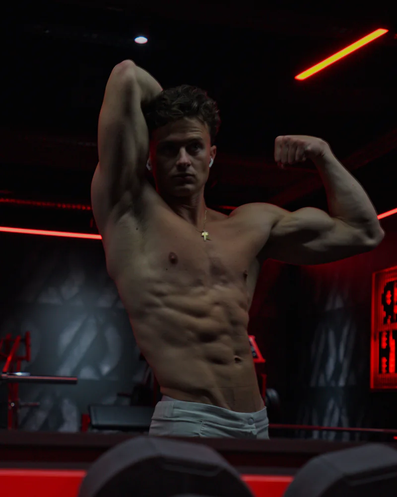 Paul, torse nu dans une salle de sport aux néons rouges, un bras levé derrière la tête, l'autre fléchi.